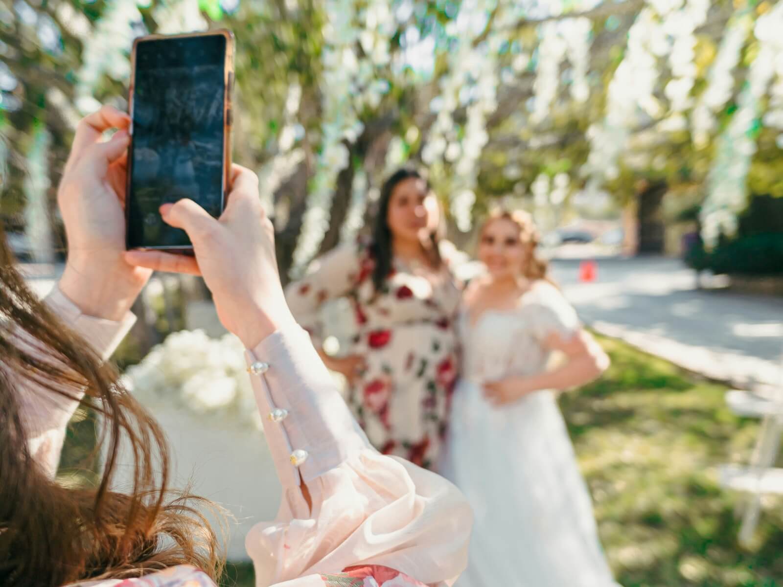 Hochzeitsgast fotografiert die Braut mit dem Smartphone im Freien.